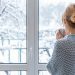 A woman in a cozy sweater stands at the picture window with a cup of drink in her hands